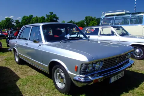 Srebrny Fiat 130 z lat 70. prezentuje się elegancko na trawie, obok innych klasyków motoryzacji.