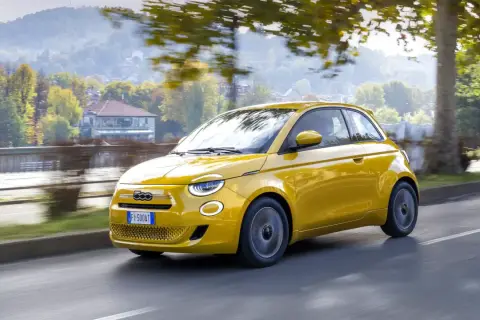 Nowy Fiat 500 w kolorze żółtym sunie po drodze, otoczony jesienną zielenią i drzewami.