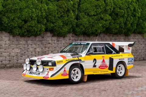 Biało-żółty Audi Sport Quattro z numerem 2, gotowy do rajdu, z licznymi światłami i logotypami sponsorów.