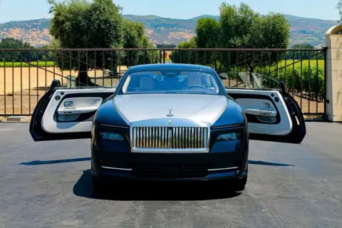 Elegancki Rolls-Royce Spectre z otwartymi drzwiami, zaparkowany przed bramą posiadłości z widokiem na wzgórza.