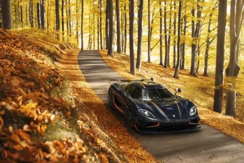 Mroczny Koenigsegg Agera RS z pomarańczowymi akcentami przemierza jesienną drogę wśród złotych liści i drzew.