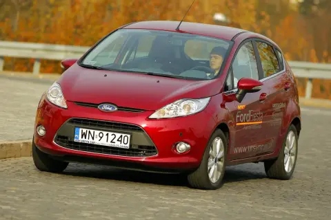Czerwony Ford Fiesta 2010 z kierowcą za kierownicą, jadący po drodze.