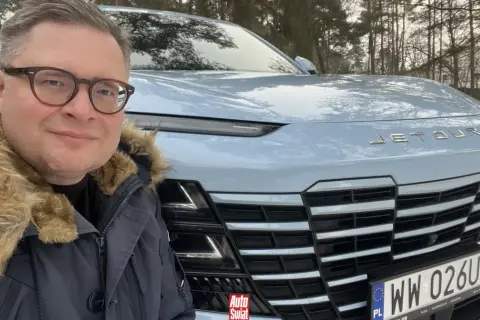 Mężczyzna w okularach i futrzanej kurtce pozuje do selfie przed jasnoniebieskim Jetour Dashing na tle zimowego lasu.