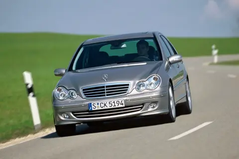 Srebrny Mercedes W203 1.8 Kompressor z kierowcą za kierownicą jedzie po krętej drodze wśród zielonych wzgórz.