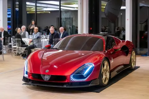 Lśniący, czerwony Alfa Romeo 33 z złotymi felgami prezentuje się na tle sali bankietowej, gdzie goście przyglądają się mu z zaciekawieniem.