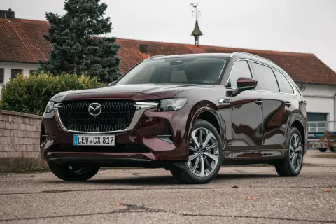 Elegancka, ciemnobordowa Mazda CX-80 stoi przed budynkiem z czerwoną dachówką.