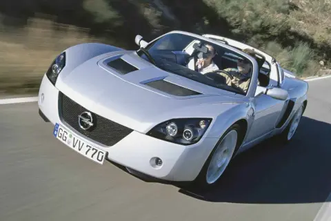 Srebrny Opel Speedster z parą na pokładzie mknie po krętej drodze, ciesząc się słońcem i wolnością.
