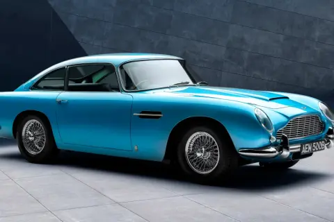 Błękitny Aston Martin DB5 z charakterystycznymi szprychowymi felgami, lśniącym chromem i elegancką linią nadwozia, zaparkowany przed nowoczesną, betonową ścianą.
