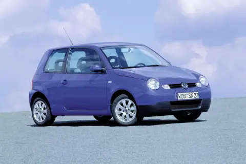 Fioletowy Volkswagen Lupo z tablicą rejestracyjną WOB SN 23 stoi na szarej nawierzchni pod błękitnym niebem z chmurami.