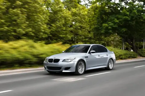 Srebrne BMW M5 V10 pędzi po drodze, otoczone zielenią drzew i krzewów.