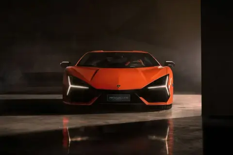Pomarańczowy Lamborghini Revuelto z ostrymi światłami Y, stojący na błyszczącej podłodze w ciemnym pomieszczeniu.