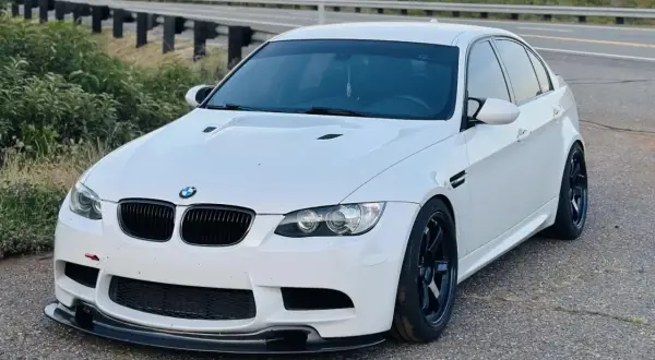Białe BMW E90 M3 z czarnymi felgami i spoilerem, zaparkowane przy drodze z barierkami.