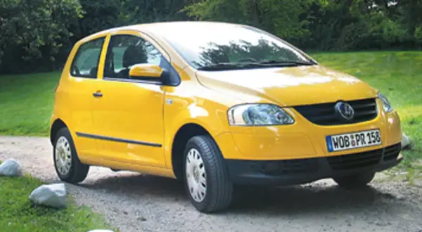 Żółty Volkswagen Fox zaparkowany na żwirowej drodze, otoczony zielenią.