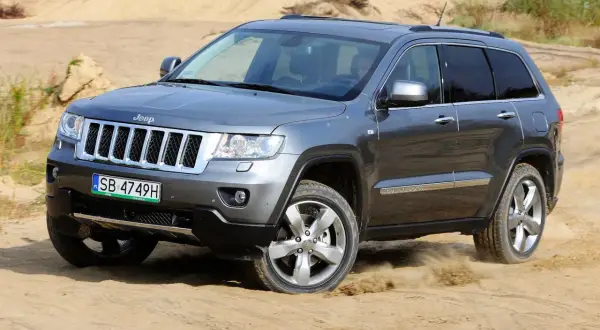 Szary Jeep Grand Cherokee 3.6 pokonuje piaszczysty teren, wzbijając tumany kurzu.
