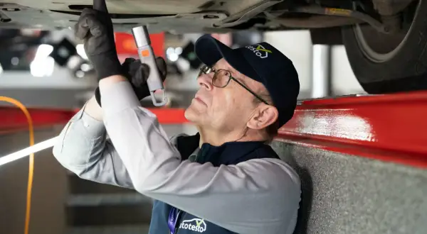 Mechanik w czapce z logo "autotesto.pl" sprawdza podwozie auta, szukając usterek. To kluczowe dla rankingu najlepszych firm od sprawdzania auta przed zakupem.