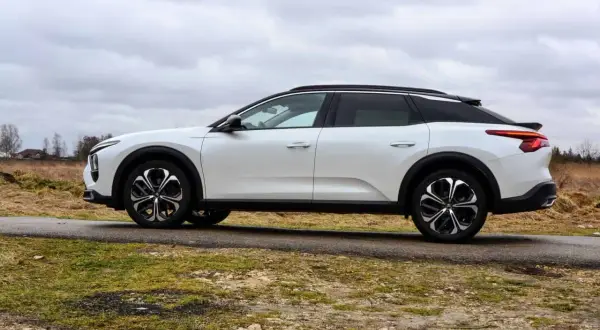 Biały Citroen C5X na tle pochmurnego nieba i wiejskiego krajobrazu. Elegancki design i nowoczesne felgi podkreślają jego wyjątkowość.