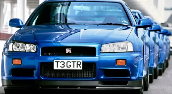 Błękitny Nissan Skyline GT-R R34 z tablicą "T3 GTR" ustawiony w rzędzie z innymi.