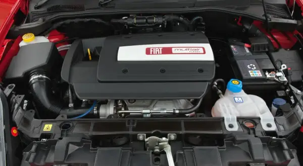 Silnik MultiAir Turbo w Fiat Grande Punto 1.4. Widoczny jest czarny plastikowy dekiel z logo Fiata, akumulator i zbiornik płynu chłodniczego.