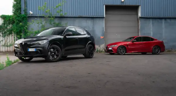 Dwa sportowe Alfa Romeo: czarny Stelvio Quadrifoglio i czerwona Giulia, stoją przed budynkiem z niebieską, falistą fasadą.