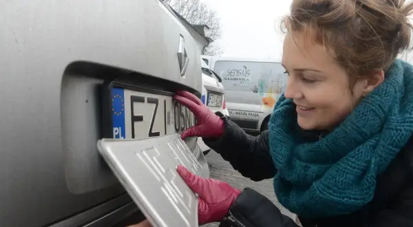 Kobieta w czerwonych rękawiczkach montuje tablicę rejestracyjną. KSU rejestracja w toku.