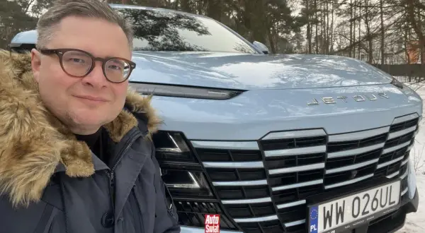 Mężczyzna w okularach i futrzanej kurtce pozuje do selfie przed jasnoniebieskim Jetour Dashing na tle zimowego lasu.