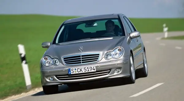 Srebrny Mercedes W203 1.8 Kompressor z kierowcą za kierownicą jedzie po krętej drodze wśród zielonych wzgórz.