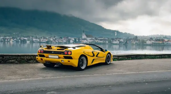 Żółty Lamborghini Diablo SV z czarnymi akcentami stoi nad jeziorem, w tle miasteczko i góry.