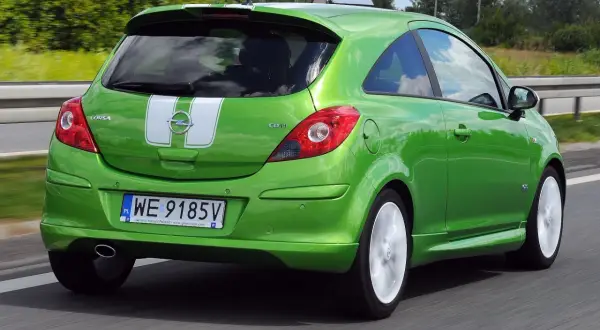 Zielony Opel Corsa D 1.2 benzyna z białymi pasami na klapie bagażnika jedzie po drodze.