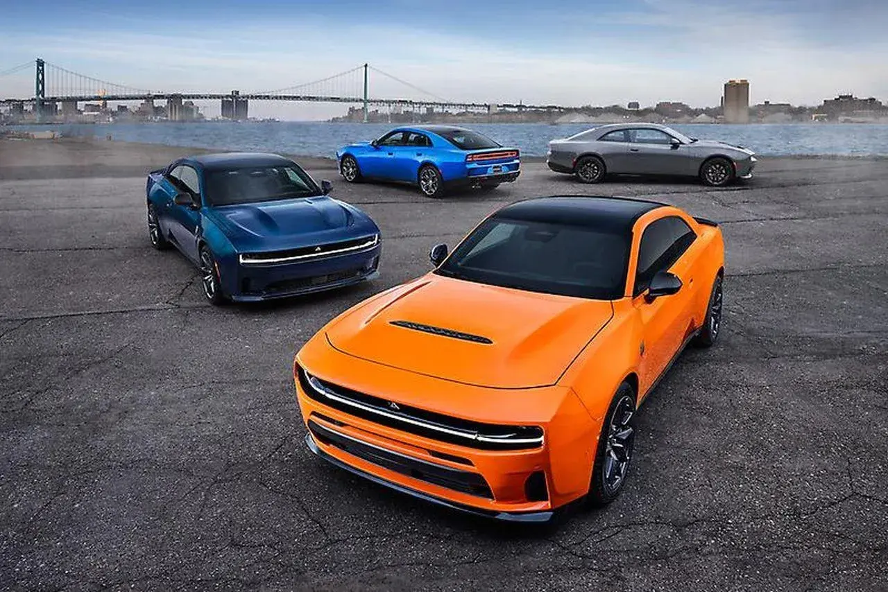 Pomarańczowy i niebieski Dodge Charger, szary Dodge Challenger i niebieski Dodge Charger stoją nad wodą z mostem w tle.