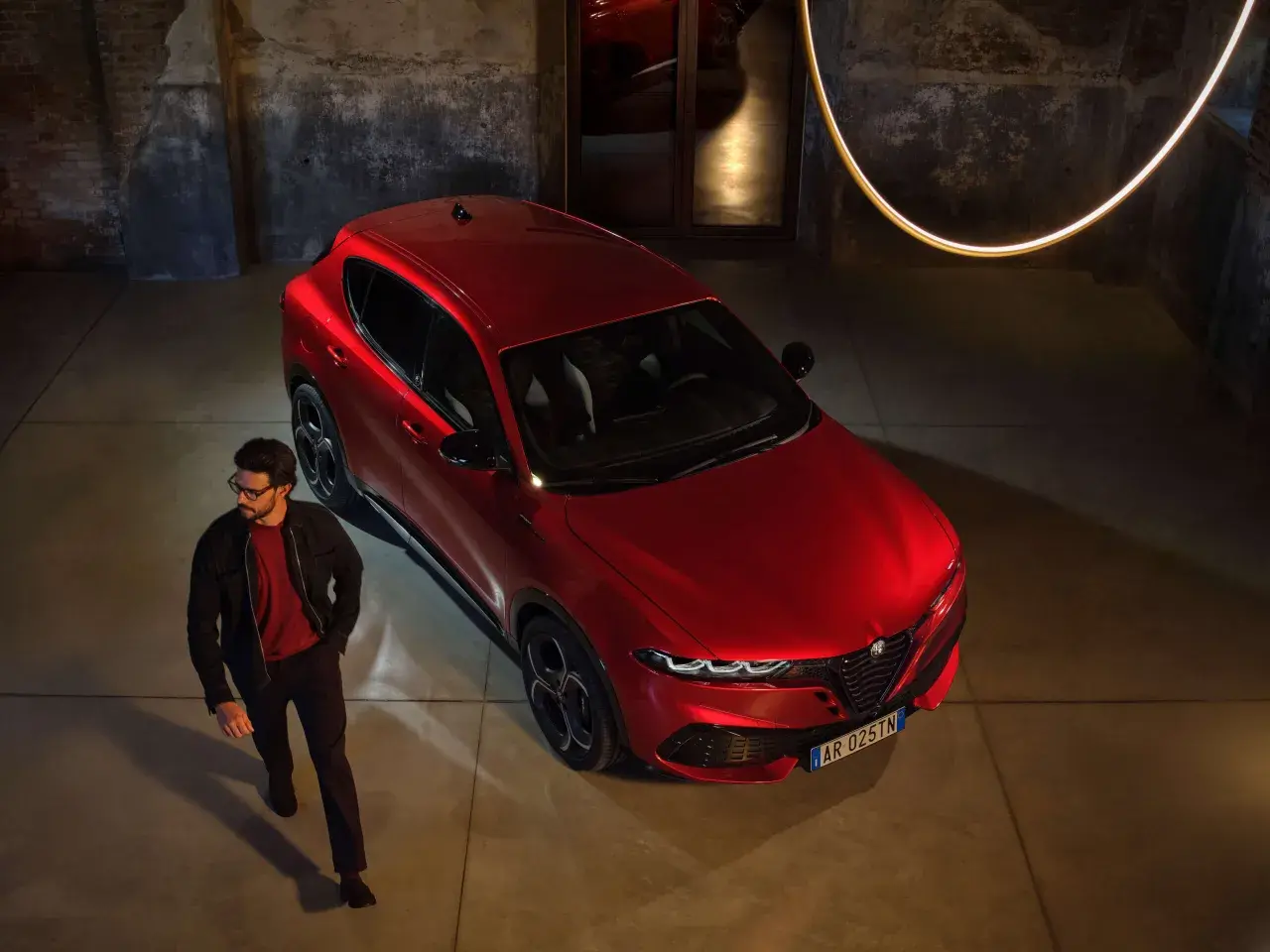 Elegancki mężczyzna w okularach obok lśniącego, czerwonego Alfa Romeo Tonale. Stylowy duet w industrialnym wnętrzu.