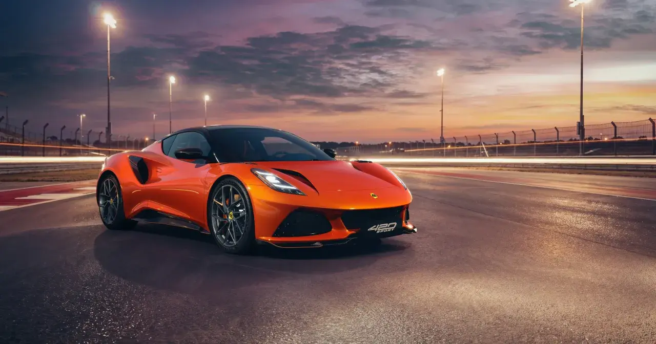 Pomarańczowy Lotus Emira 420 Sport na torze wyścigowym o zachodzie słońca, z rozmazanymi światłami w tle.