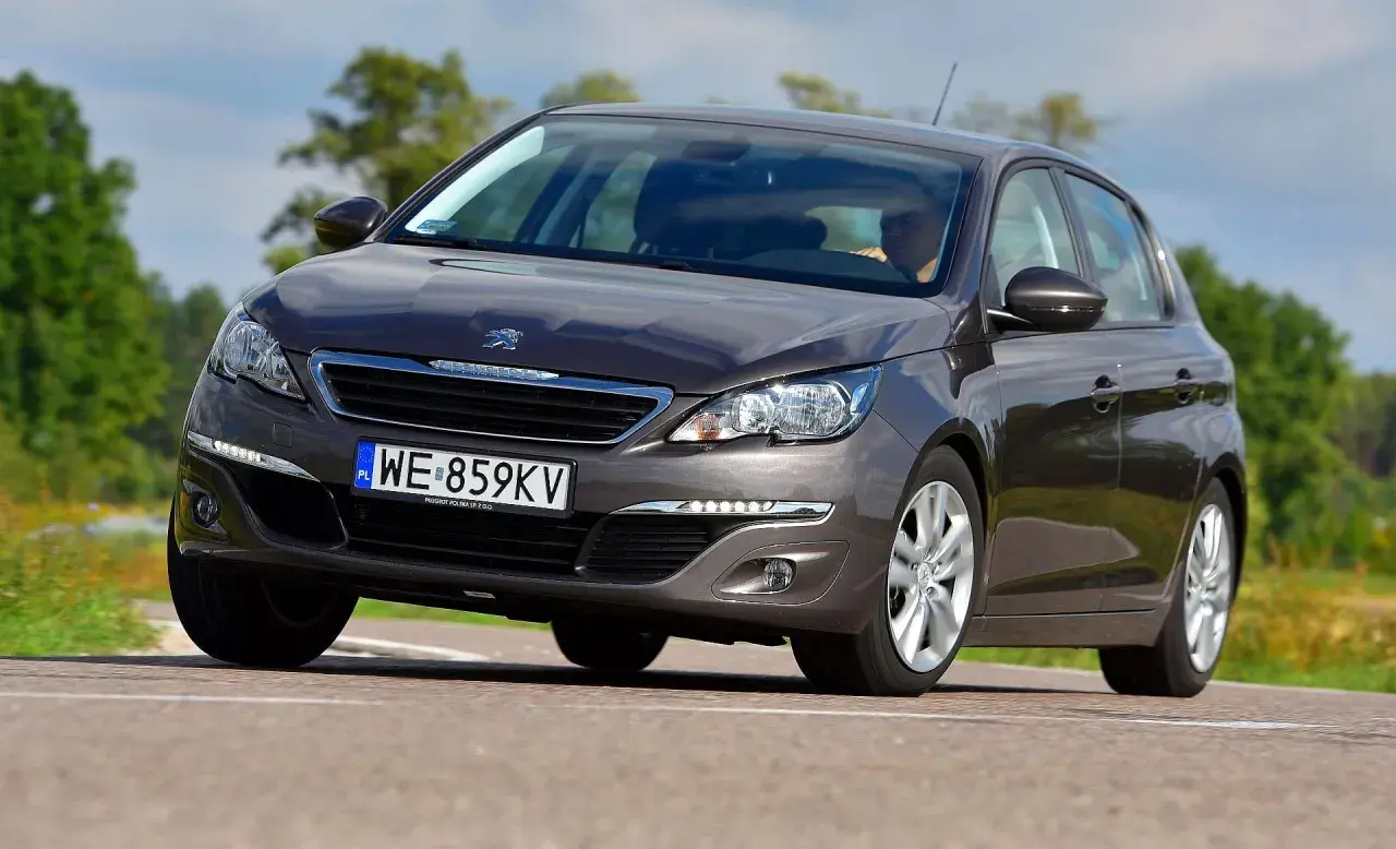 Ciemnoszary Peugeot 308 2015 z polskimi tablicami WE 859KV jedzie po drodze wśród zieleni.