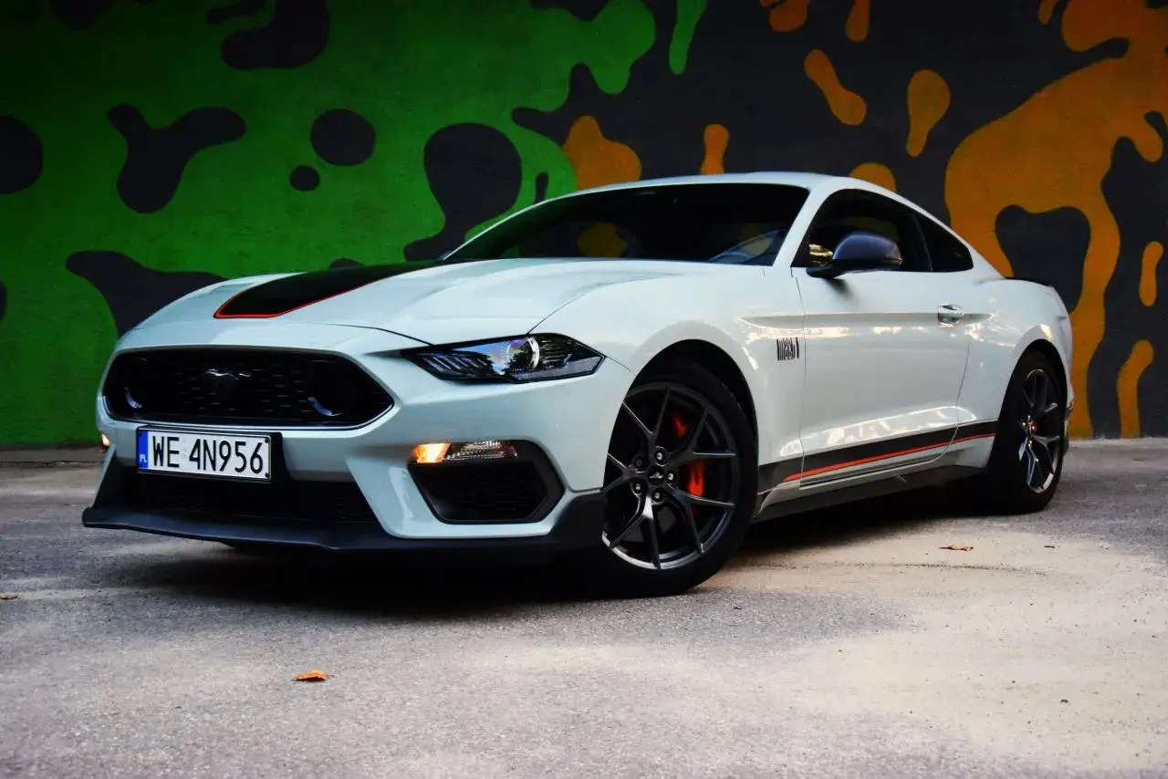 Biały Ford Mustang Mach 1 z czarnymi pasami i pomarańczowymi akcentami na tle kolorowego muralu.