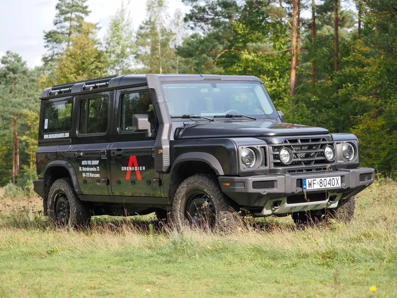 Czarny INEOS Grenadier z logo "A" na drzwiach, zaparkowany na trawie, z lasem w tle.