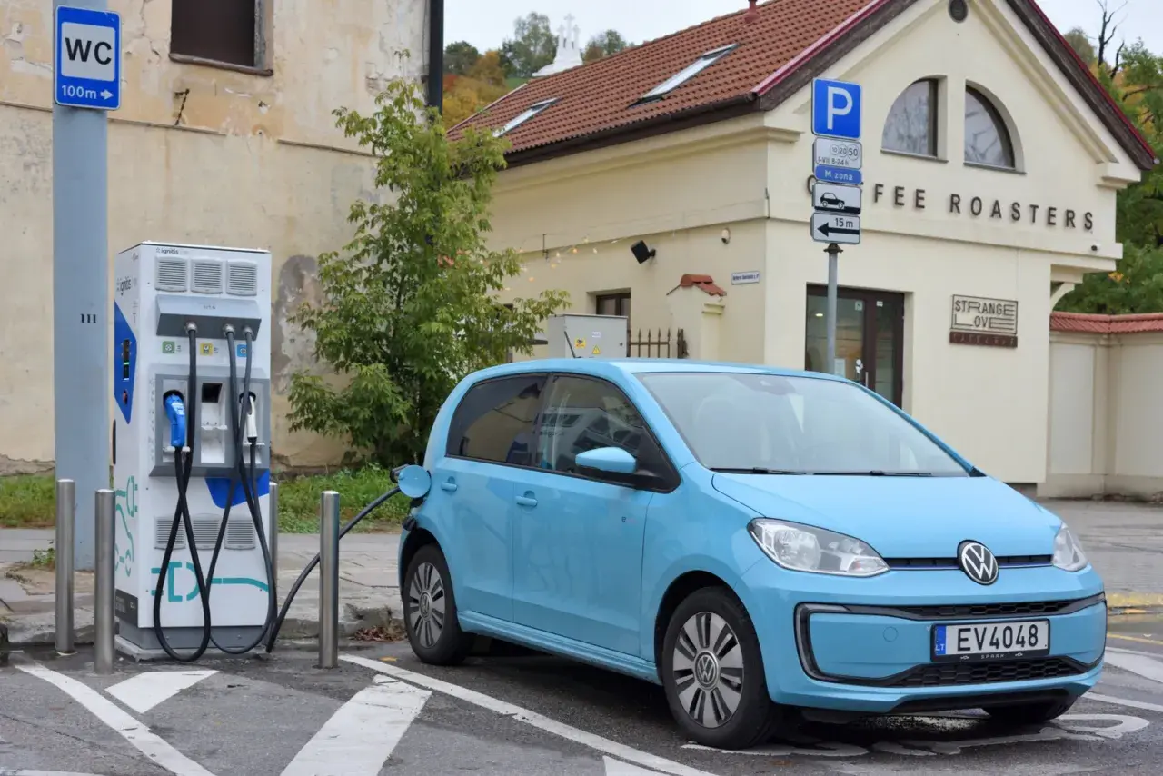 Niebieski Volkswagen Up ładuje się przy stacji ładowania. Obok znajduje się budynek z napisem "COFFEE ROASTERS".