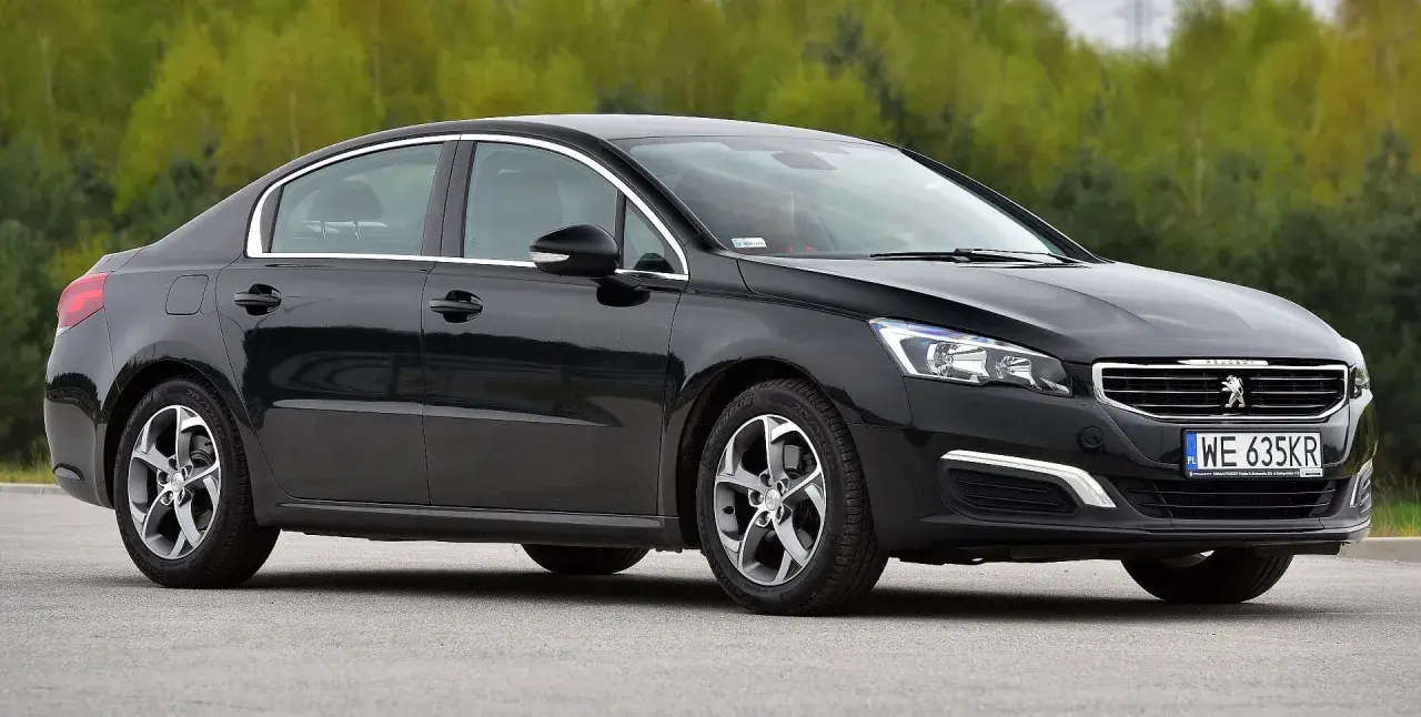 Elegancki, czarny Peugeot 508 z chromowanymi detalami i felgami stoi na tle zieleni.