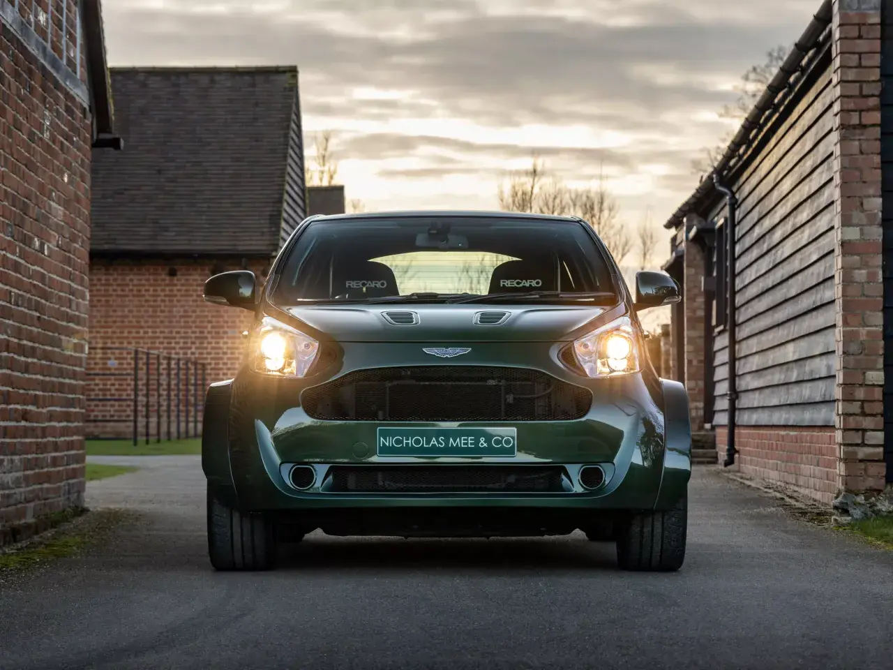 Aston Martin Cygnet w ciemnozielonym kolorze, z reflektorami świecącymi w stronę widza, zaparkowany na podjeździe między ceglanymi budynkami.
