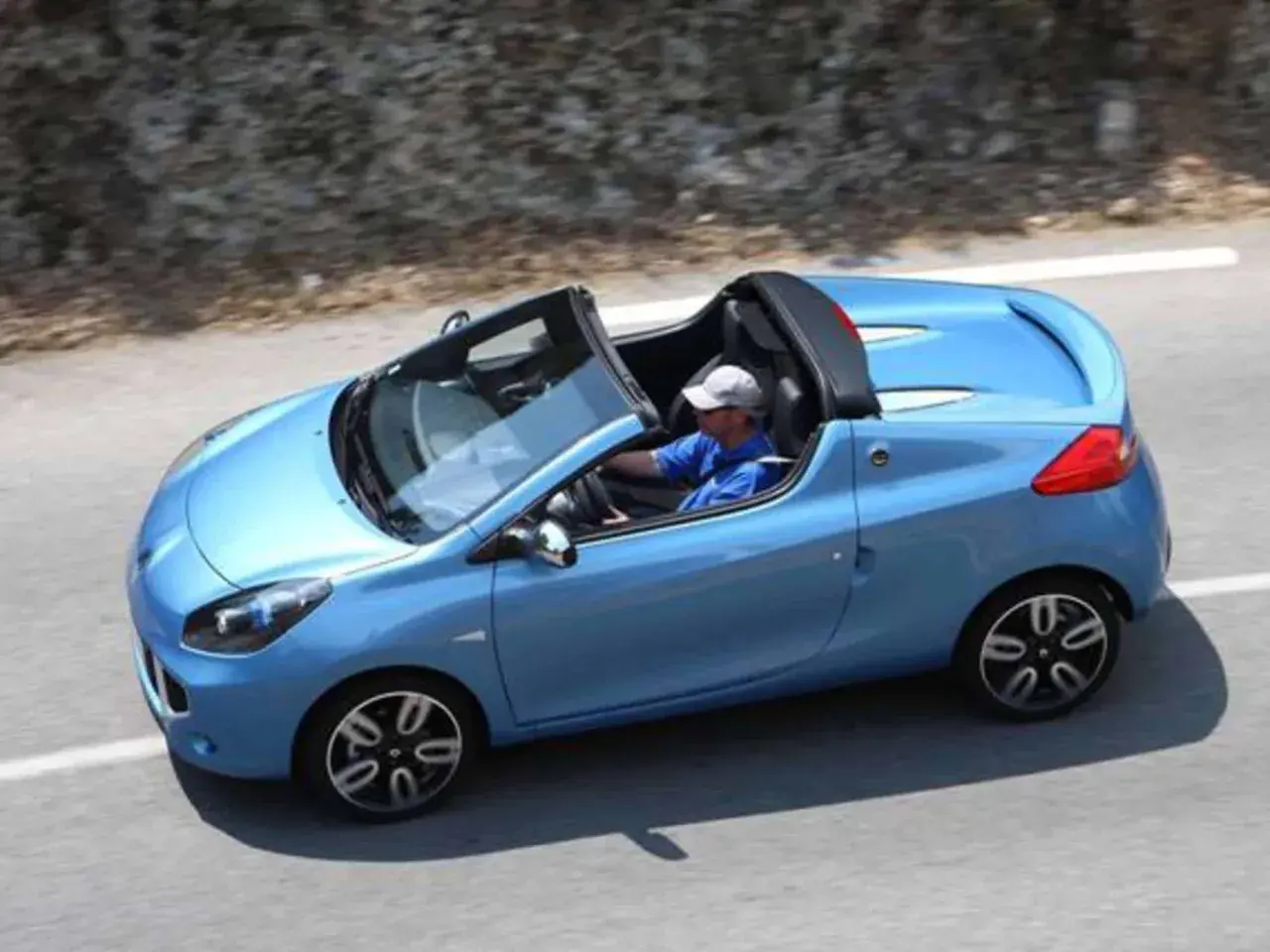 Niebieski kabriolet Renault Wind z otwartym dachem, kierowca w czapce i niebieskiej koszulce jedzie po krętej drodze.