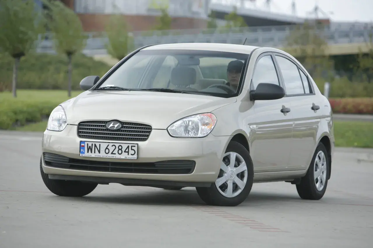 Złoty Hyundai Accent z kierowcą za kierownicą, zaparkowany na zewnątrz.