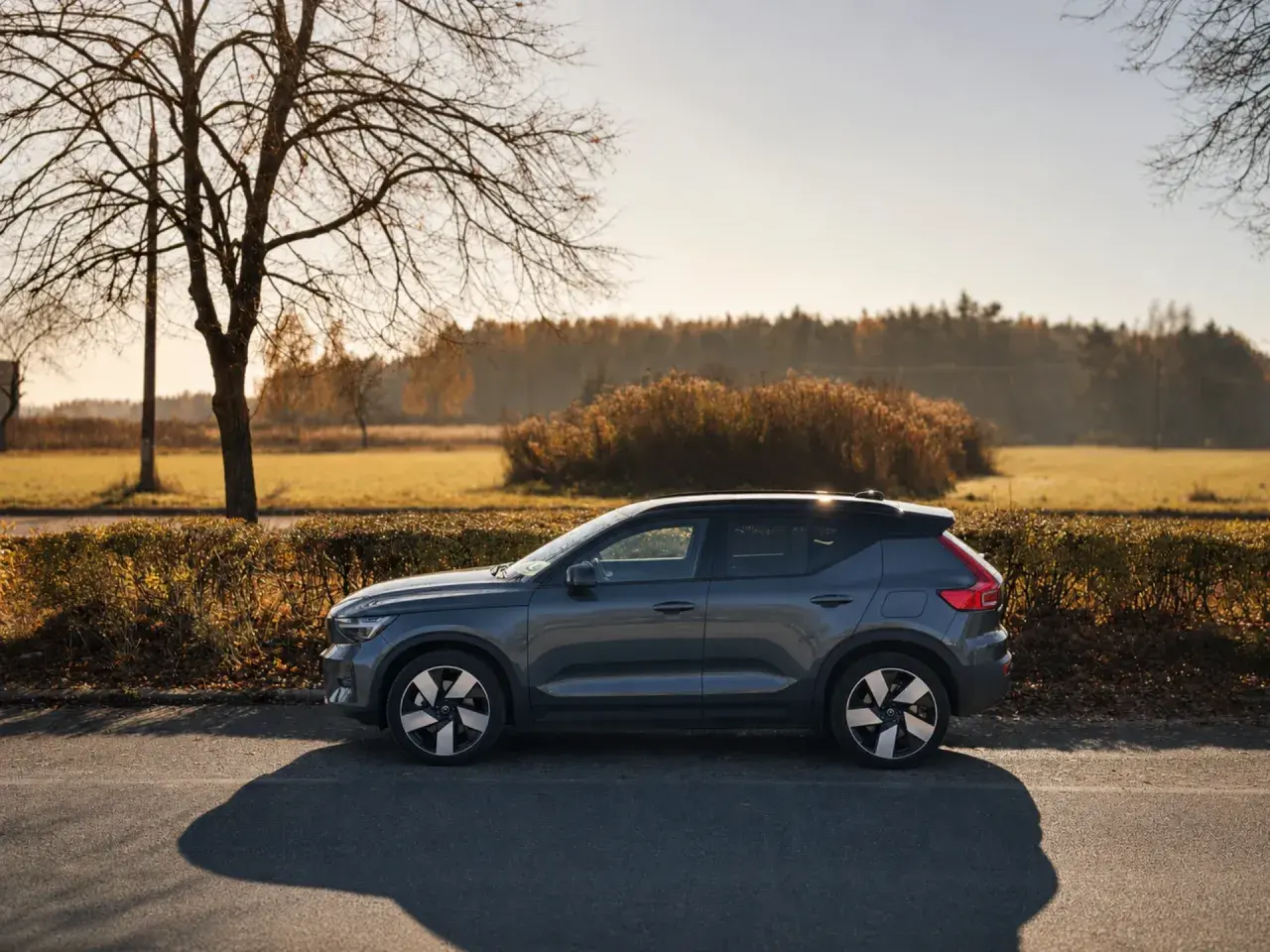 Szary Volvo EX40 zaparkowany na poboczu drogi, w tle jesienny krajobraz z drzewami i krzewami.