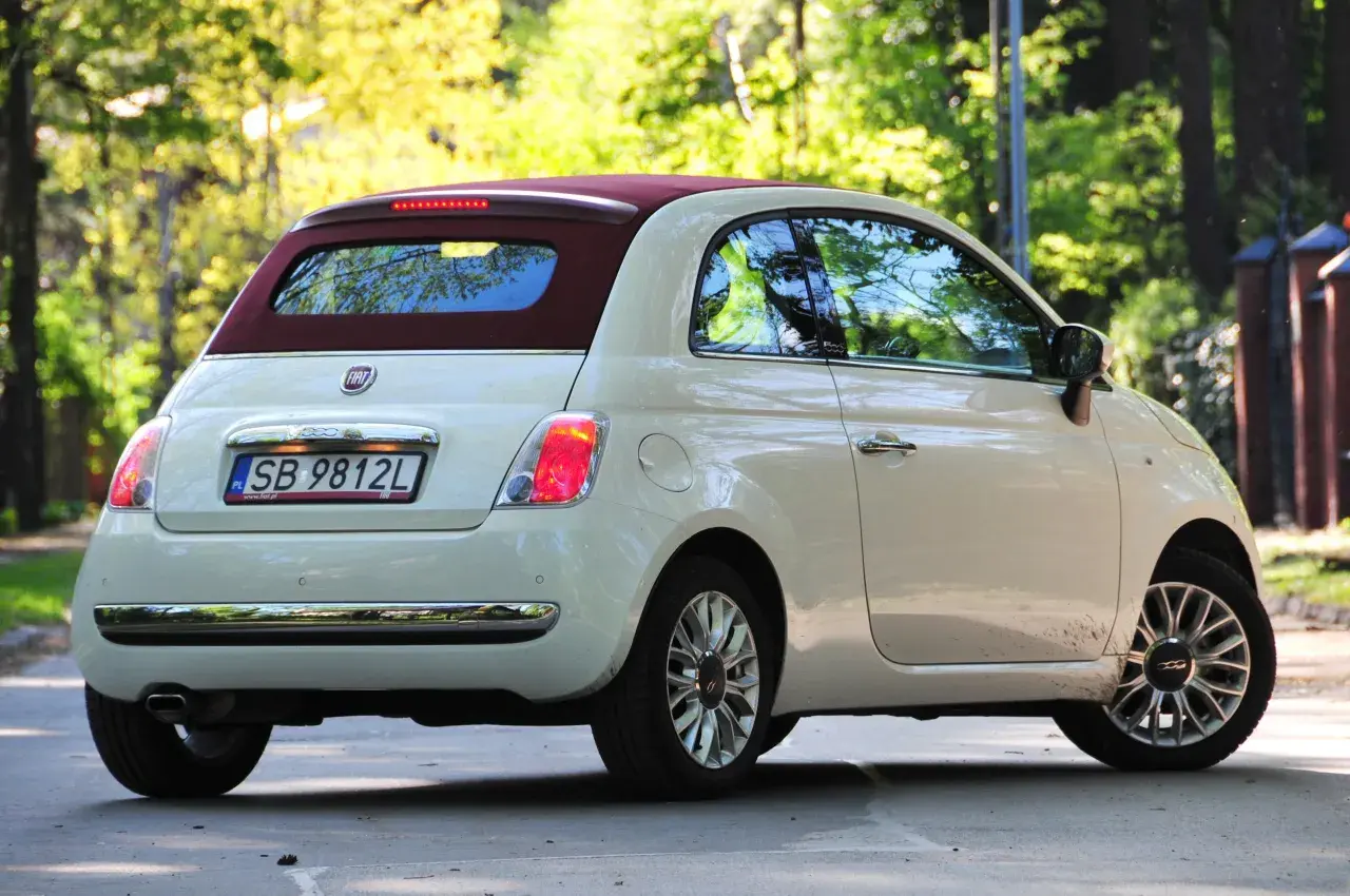 Biały fiat 500c z bordowym dachem stoi na drodze, otoczony zielenią drzew.