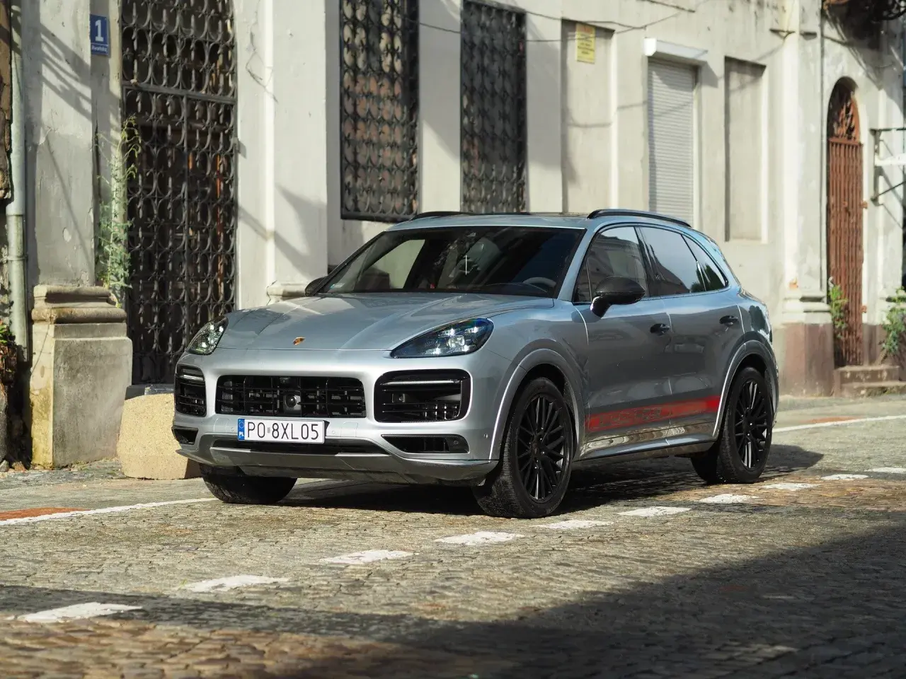 Srebrny Porsche Cayenne GTS z czerwonymi akcentami na progach, zaparkowany na brukowanej ulicy przed zabytkową kamienicą.