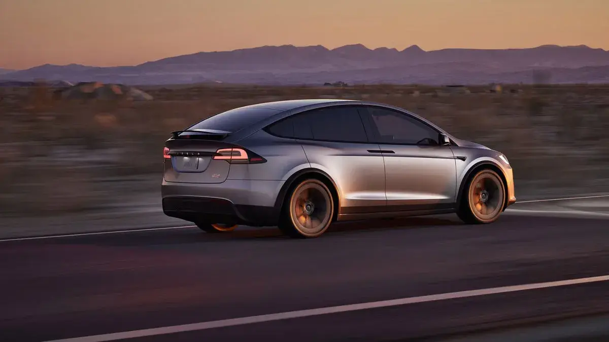Srebrny Tesla X pędzi przez pustynię o zachodzie słońca, góry w tle.