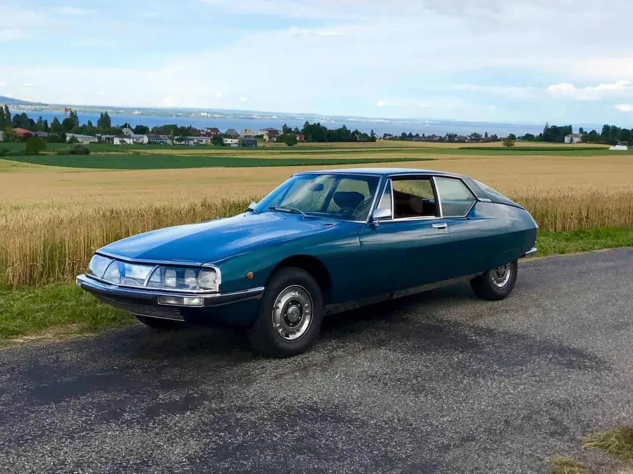 Piękny, turkusowy Citroen SM na tle złotego pola i jeziora. Klasyk motoryzacji w malowniczej scenerii.