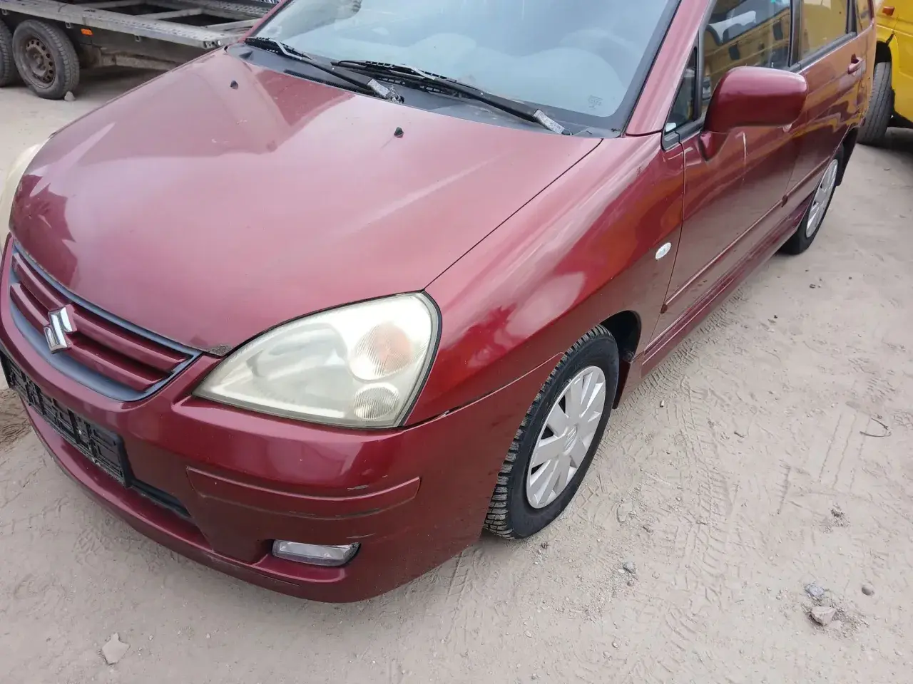 Czerwony Suzuki Liana stoi na piaszczystym podłożu. Widoczny przód auta z reflektorem i grillem.