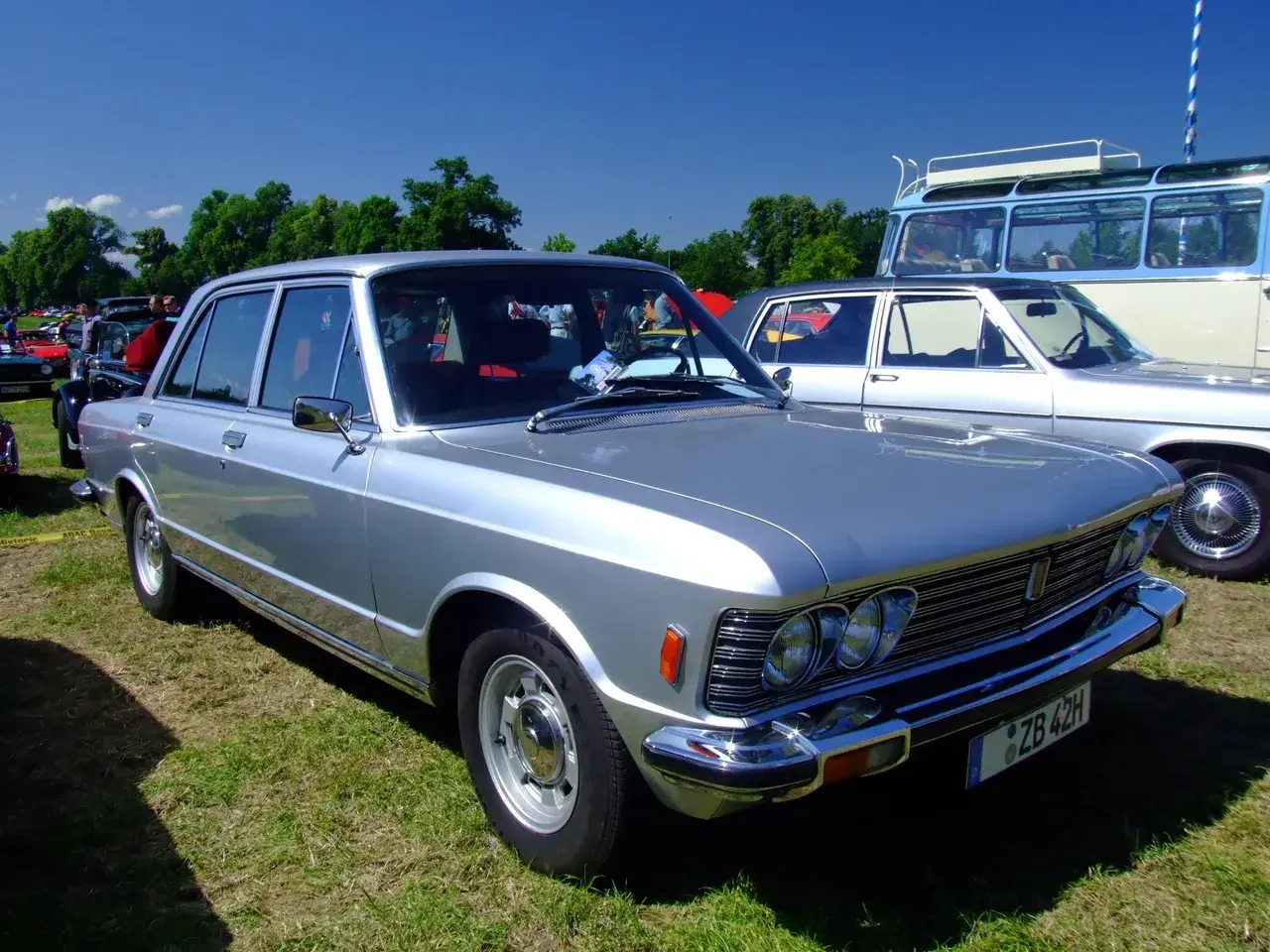 Srebrny Fiat 130 z lat 70. prezentuje się elegancko na trawie, obok innych klasyków motoryzacji.