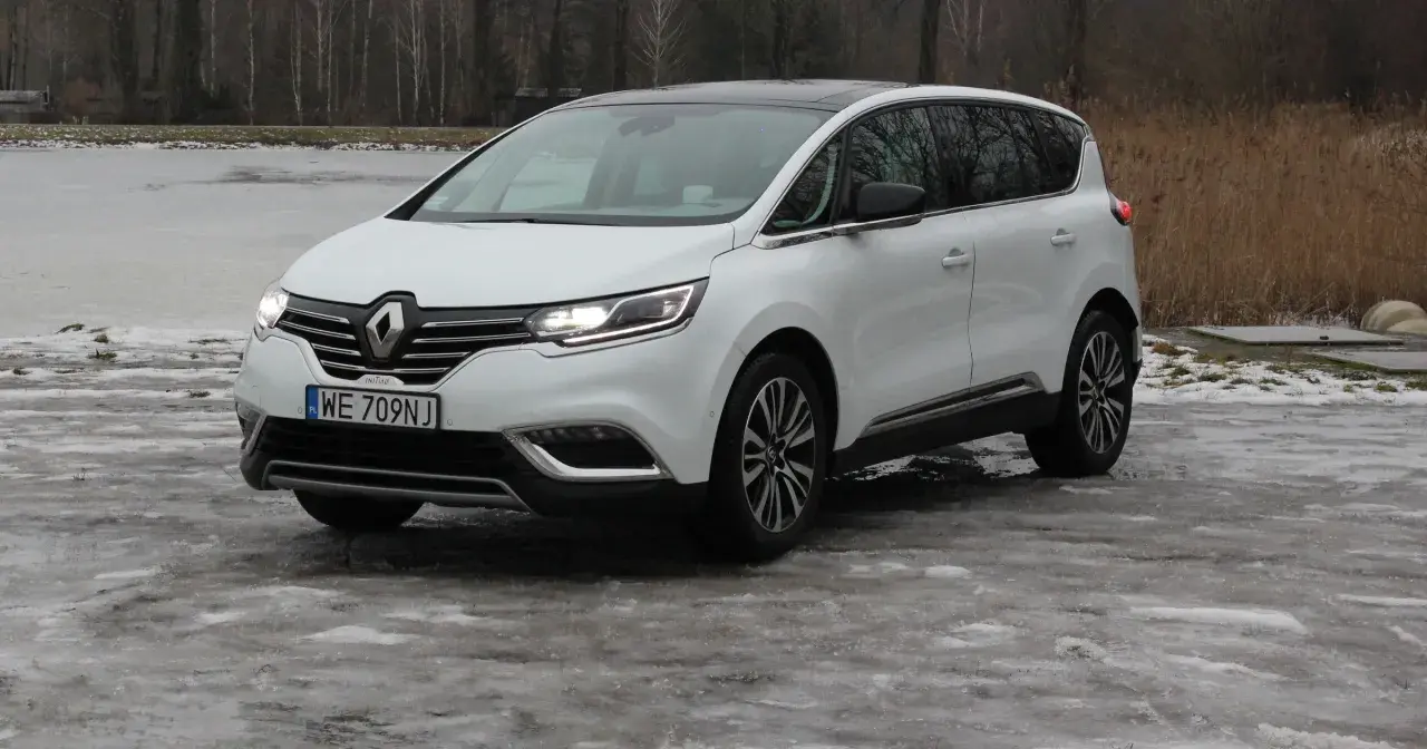 Biały Renault Initiale na oblodzonym podjeździe, z widokiem na zamarznięte jezioro i drzewa.