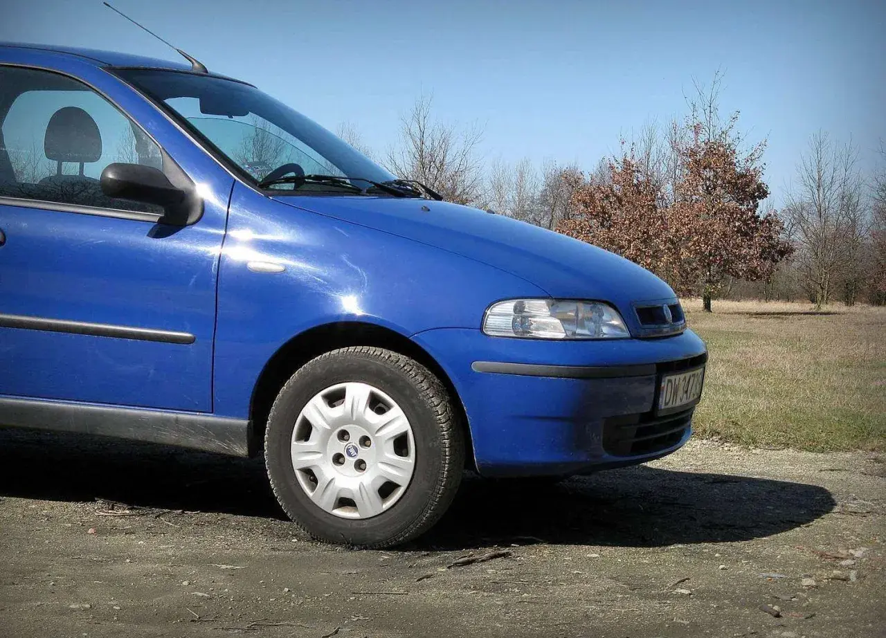 Błękitny Fiat Albea stoi na polnej drodze, w tle jesienne drzewa.