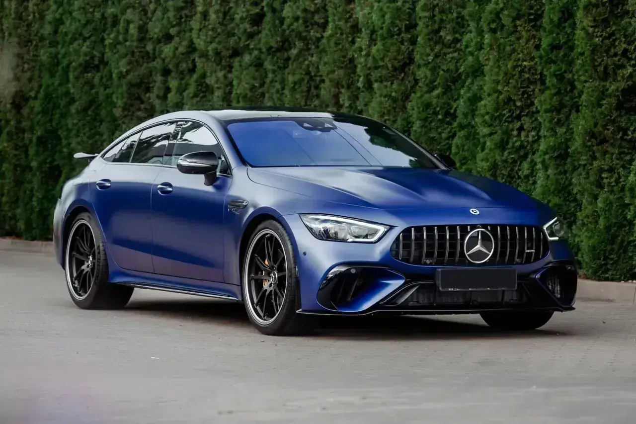 Matowy, granatowy Mercedes E63 AMG z czarnymi felgami stoi na tle zielonego żywopłotu.