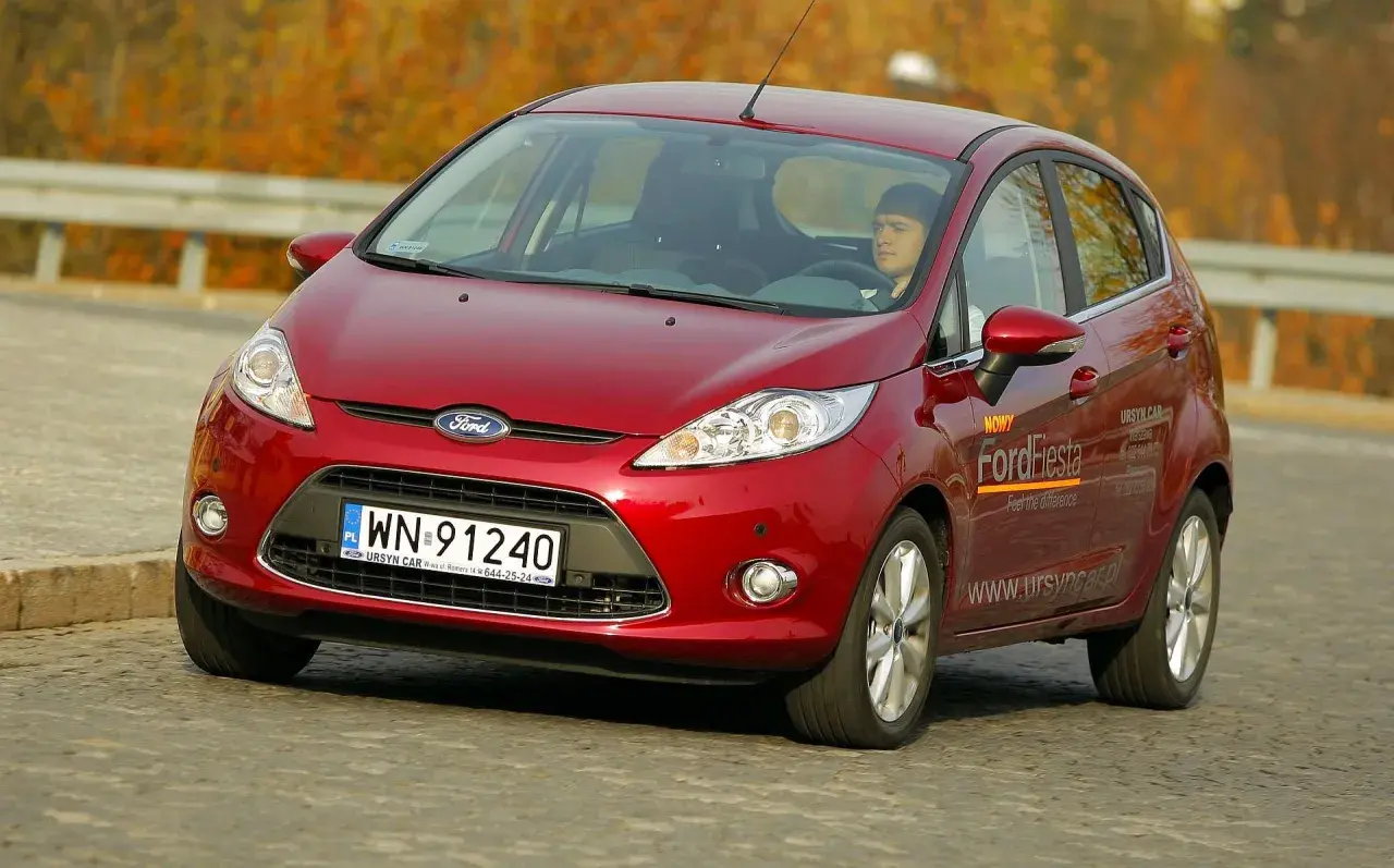 Czerwony Ford Fiesta 2010 z kierowcą za kierownicą, jadący po drodze.